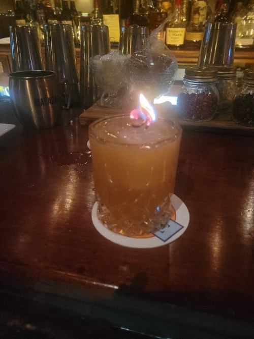A cocktail with a flaming garnish sits on a wooden bar counter, surrounded by various bar tools and bottles.