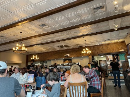 A busy restaurant interior with diners at tables, a visible kitchen area, and warm lighting from hanging fixtures.