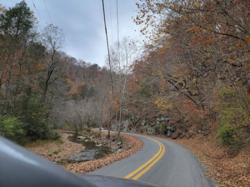 A winding road surrounded by autumn trees and a creek, with gray skies overhead.