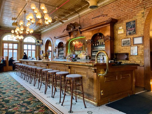 A vintage bar with wooden decor, high stools, and a well-stocked counter, illuminated by warm lighting and chandeliers.