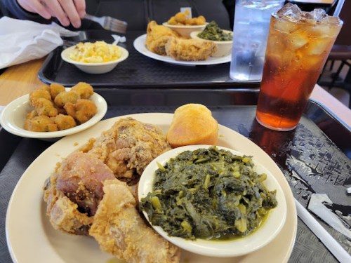 A plate of fried chicken, collard greens, cornbread, and okra, with a glass of iced tea and a side of macaroni and cheese.