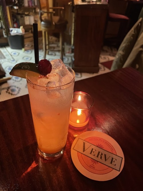 A refreshing cocktail with ice, a lime wedge, and a cherry, next to a lit candle and a coaster labeled "VERVE."