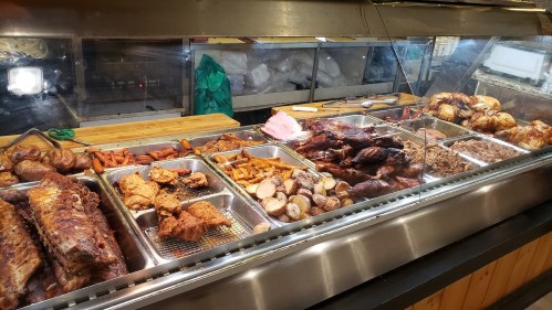 A variety of cooked meats and sides displayed in metal trays, including ribs, chicken, and potatoes.