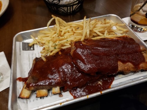 A metal tray holds BBQ ribs covered in sauce, accompanied by a side of crispy French fries.