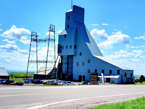 A large blue industrial building with a tall structure and power lines, set against a clear blue sky.