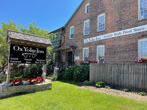 A brick building with a sign for Ox Yoke Inn, featuring flowers and greenery in a sunny outdoor setting.
