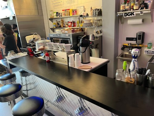 A diner counter with coffee pots, condiments, and a busy kitchen in the background. Stools line the counter.
