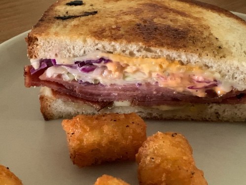A toasted sandwich with layers of ham, cheese, and coleslaw, served with crispy tater tots on the side.