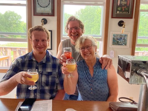 Three people smiling and holding drinks at a bar, with a bright, sunny background and framed pictures on the walls.