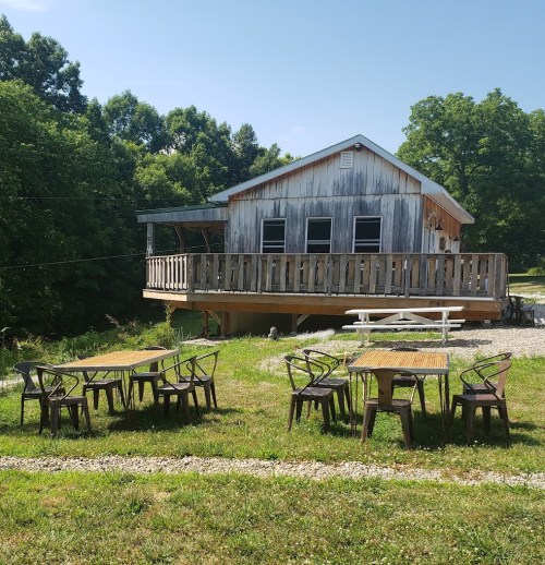 A rustic cabin with a porch, surrounded by green grass and several outdoor tables in a sunny setting.