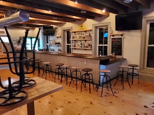 A cozy bar interior with a wooden counter, stools, and shelves of liquor, featuring exposed beams and warm lighting.