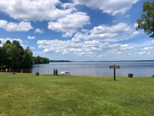 A serene lakeside view with green grass, trees, and fluffy clouds reflecting in the water under a blue sky.