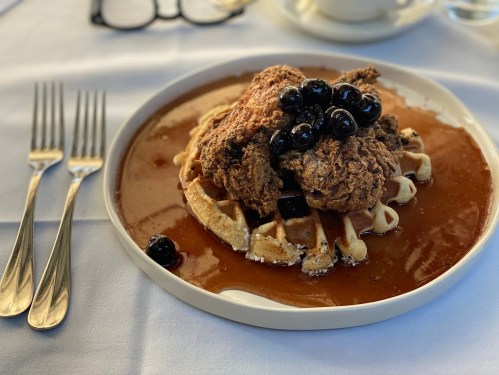 A plate of chicken and waffles topped with blueberries and syrup, set on a white tablecloth with utensils nearby.