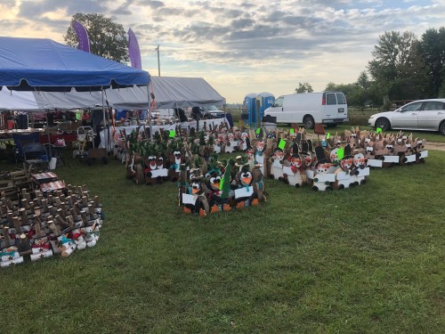 A grassy area with colorful wooden duck decorations for sale, tents in the background, and vehicles parked nearby.