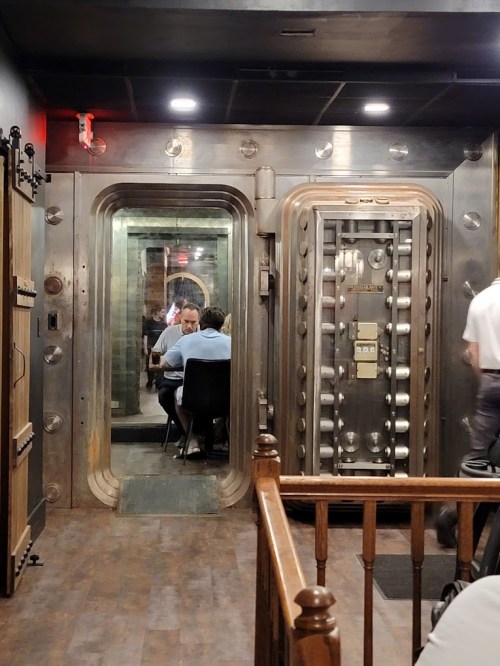 A dimly lit restaurant featuring a large, metallic vault door and a dining area visible through the entrance.