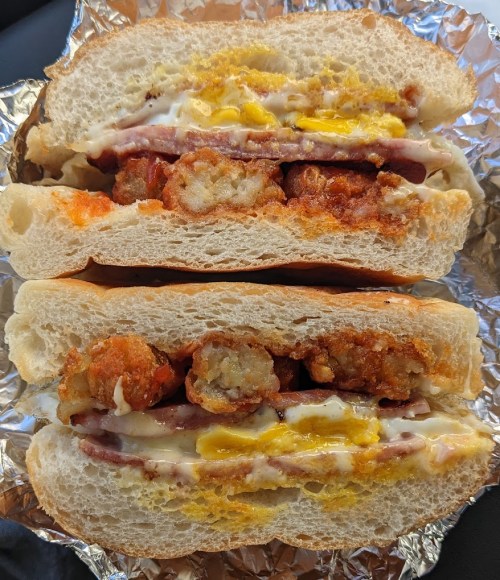 A sandwich with layers of sausage, crispy hash browns, and mustard, served on soft bread and wrapped in foil.