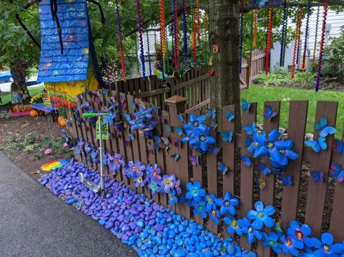 Colorful garden scene with a wooden fence adorned with blue flowers and butterflies, alongside a path of purple stones.