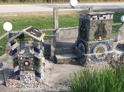 Two decorative structures made of stones and shells, surrounded by greenery, along a roadside.