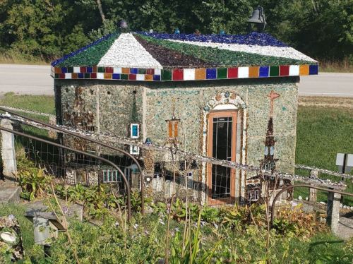 A small, uniquely designed building made of glass and stone, featuring colorful decorations and surrounded by greenery.