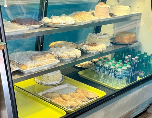 A display case filled with various cakes, pastries, and bottled water, all covered with plastic wrap.