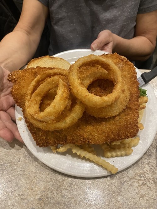 A plate with a large breaded piece of meat topped with onion rings, served with fries and a slice of bread.
