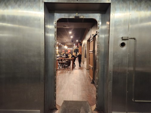 A view through a metallic doorway into a restaurant with wooden tables and a person walking inside.