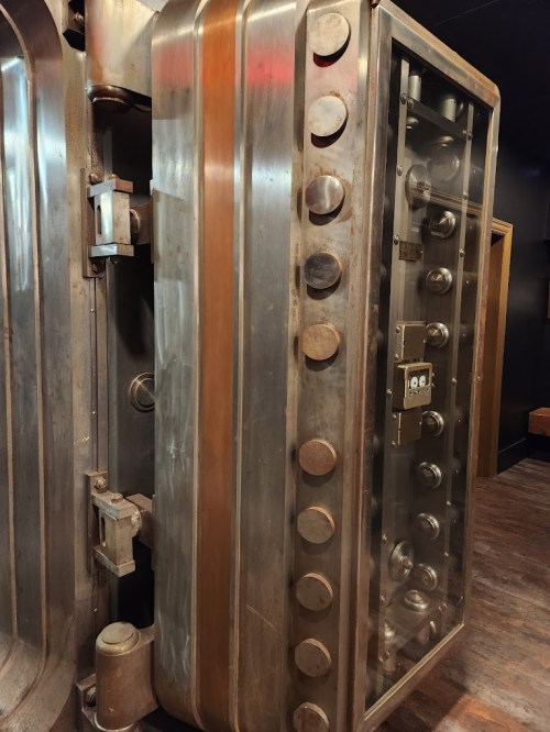 A large, metallic vault door with a glass panel and multiple locking mechanisms, set against a dark background.