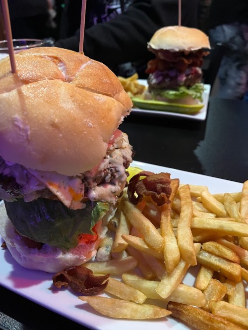 A towering burger with layers of meat, lettuce, and bacon, served with a side of golden fries on a white plate.