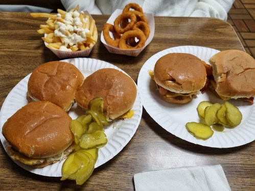 Two plates with sandwiches and pickles, accompanied by fries and onion rings in the background.