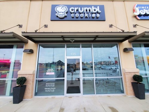Exterior of a Crumbl Cookies store, featuring large glass doors and a sign above, with planters on either side.