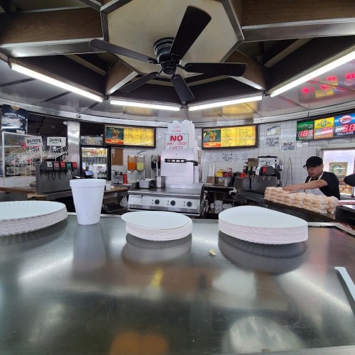 A diner counter with plates stacked, a drink cup, and a kitchen area in the background featuring a fan and menu boards.