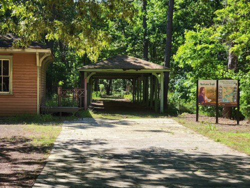 A wooden path leads through a green forest, with a gazebo and a sign for a nature trail on the side.