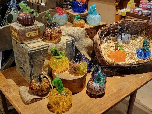 Colorful glass pumpkins in various shapes and sizes displayed on wooden crates and in a basket with straw.