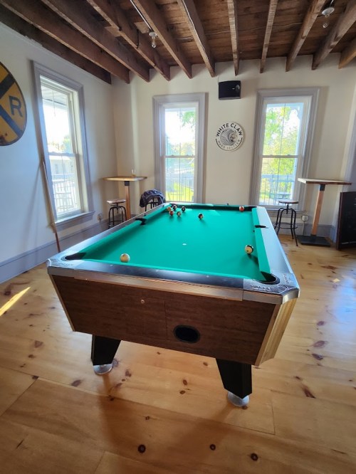A green pool table in a bright room with wooden floors and large windows, featuring a sign on the wall.