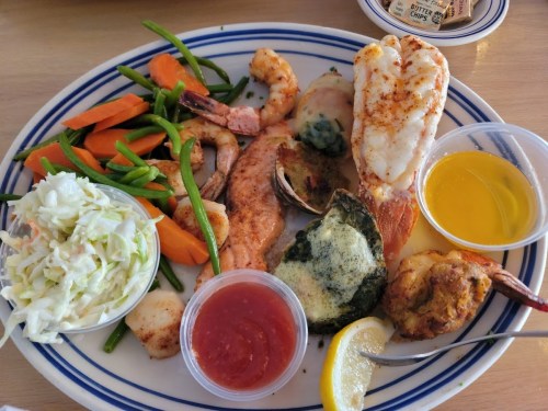 A seafood platter featuring shrimp, lobster, scallops, vegetables, coleslaw, and dipping sauces on a blue-striped plate.