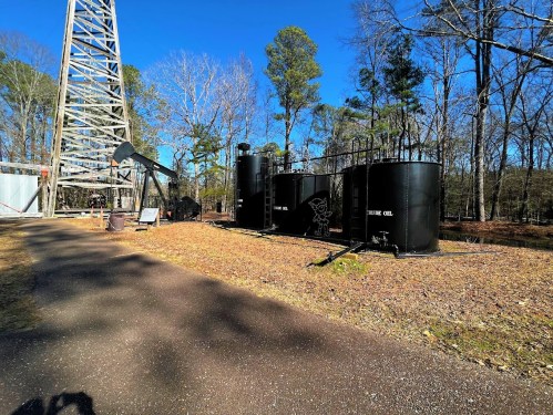 Oil storage tanks beside a pump jack and a tall structure, surrounded by trees and a clear blue sky.
