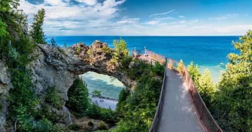 A scenic view of a rocky arch overlooking a vibrant blue lake, with a pathway and lush greenery surrounding it.