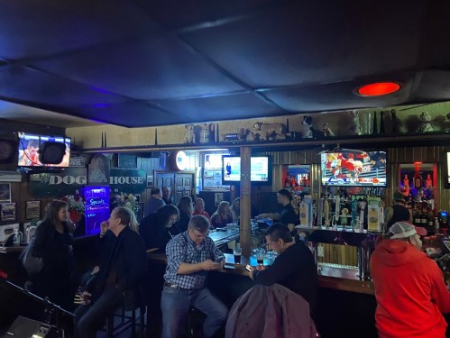 A lively bar scene with patrons at the counter, multiple TVs showing sports, and colorful lighting.