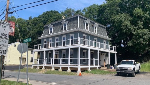 A large, two-story house with a gray exterior and a wraparound porch, located at the corner of a street.