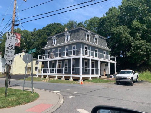 A large, two-story house with a wraparound porch at a street corner, surrounded by trees and a parked white truck.