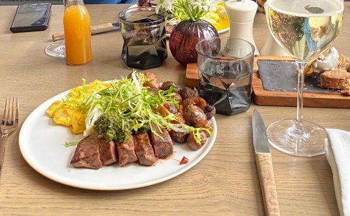 A plate with sliced steak, greens, roasted mushrooms, and an omelet, accompanied by drinks and condiments on a table.