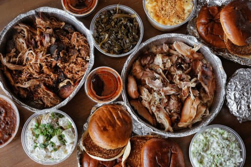 A spread of barbecue dishes including pulled pork, sides, and buns arranged on a wooden table.