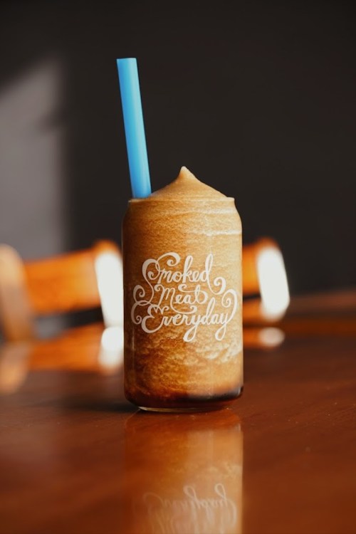 A frosty drink in a glass with a blue straw, featuring the text "Smoked Meat Everyday" on the front.