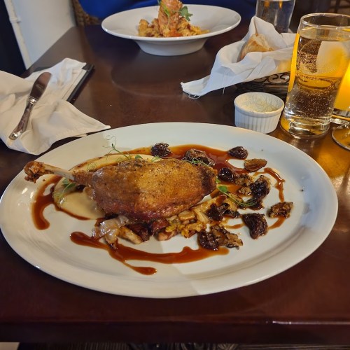 A plated dish featuring crispy duck leg, sauce, and garnishes, served on a white plate at a dining table.