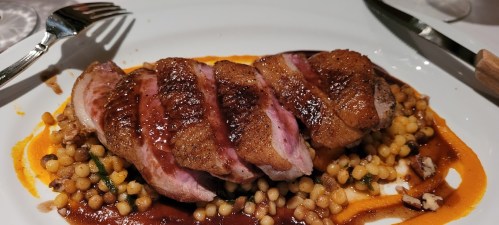 A plated dish featuring sliced duck breast on a bed of couscous, drizzled with sauce and garnished with puree.