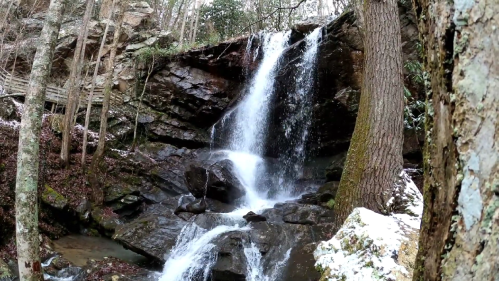 A serene waterfall cascading over rocky cliffs, surrounded by trees and a dusting of snow on the ground.