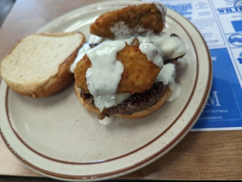 A sandwich with a beef patty, topped with crispy fried items and drizzled with white sauce, served on a plate.