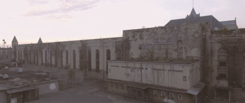 Aerial view of an abandoned, overgrown building with crumbling walls and a faded facade under a soft sunset sky.