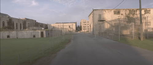 Abandoned buildings and a fenced pathway in a desolate, overgrown area under a cloudy sky.