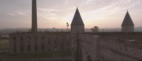 Aerial view of an old building with two spires at sunrise, featuring a smokestack in the background and a cloudy sky.